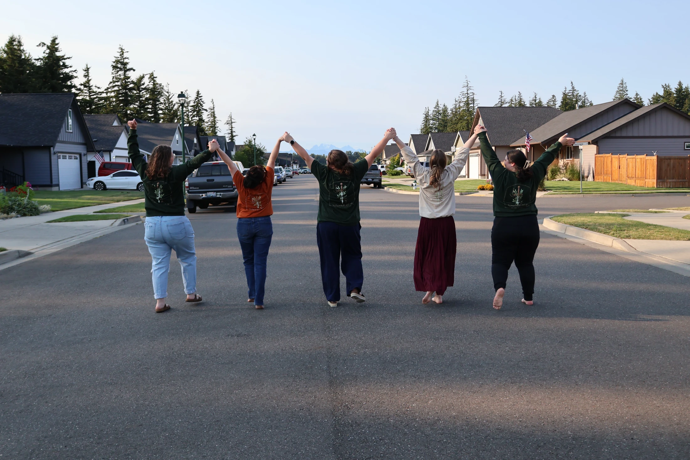 Five women seen from behind walk down the middle of a residential street, holding hands with their arms raised in a continuous chain.
