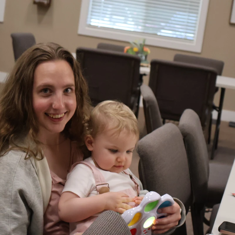 A young woman smiles at the camera while holding a small child on her lap. The child is looking down and playing with a colorful, light-up toy.