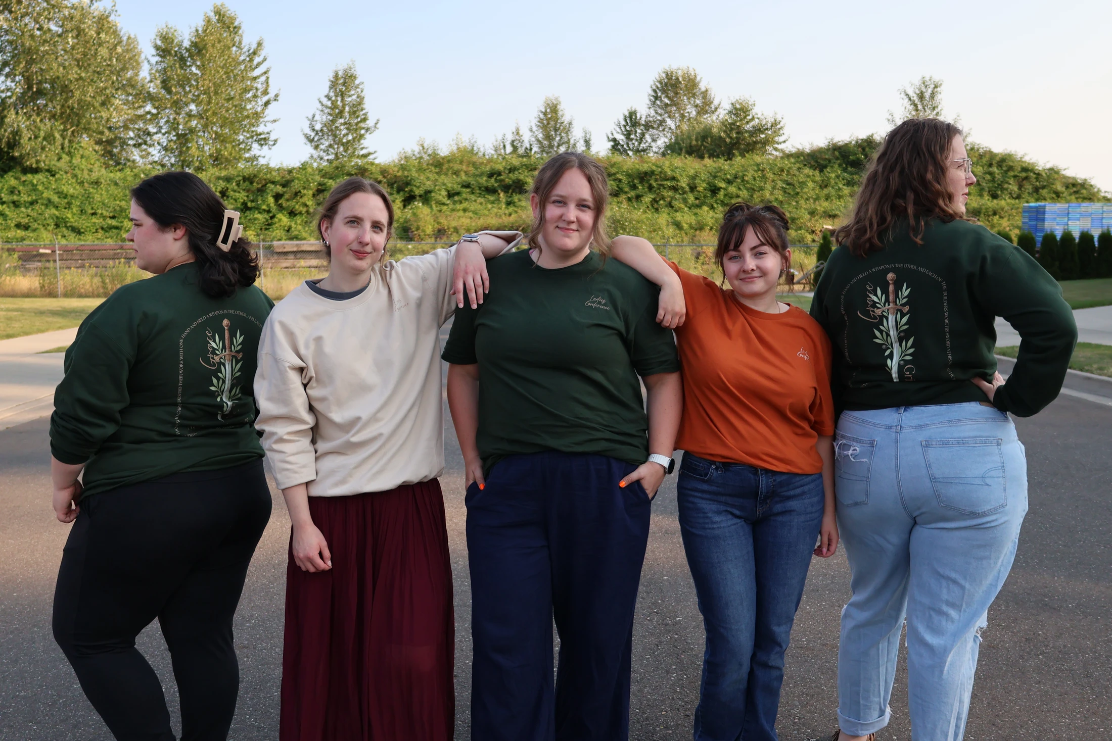A group of five young women stand side-by-side outdoors, some wearing matching green shirts from what appears to be a conference or event.