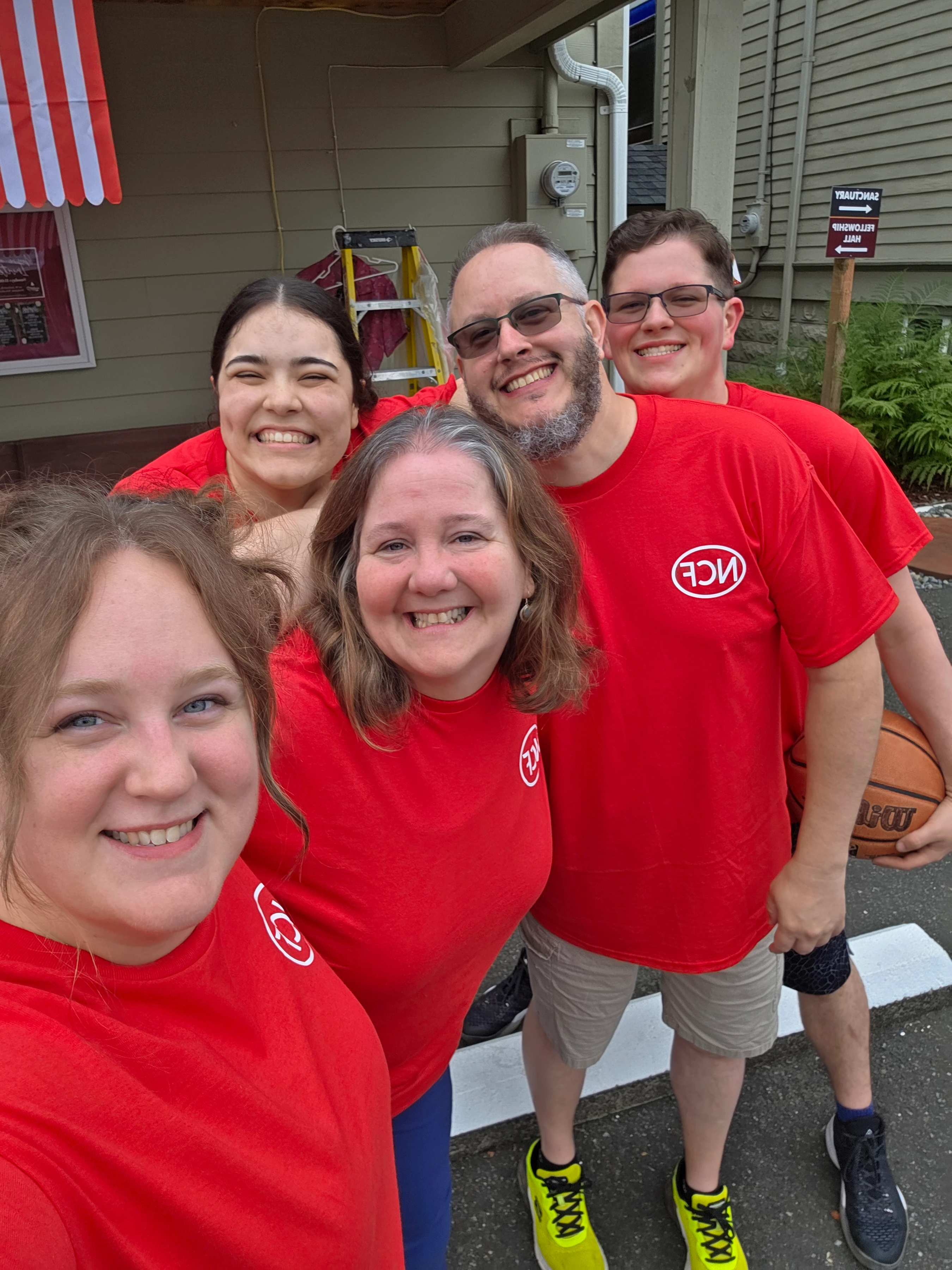 A group selfie of five smiling people, three women and two men, wearing matching red t-shirts with an 'NCF' logo, standing together outdoors.