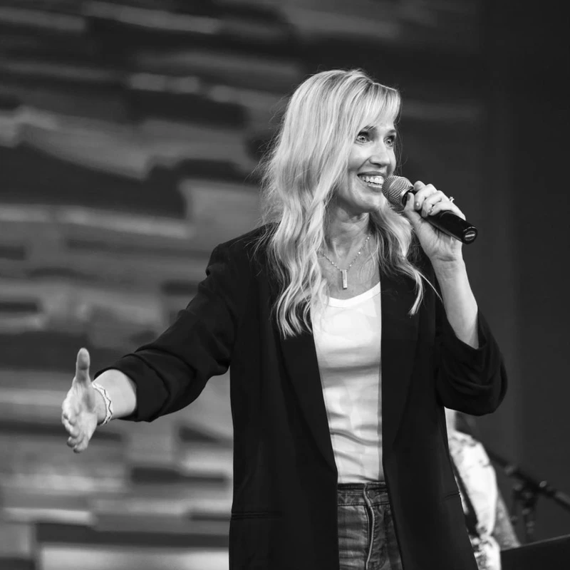 A black and white photo of a smiling blonde woman on stage. She holds a microphone and extends her arm towards the audience while speaking.