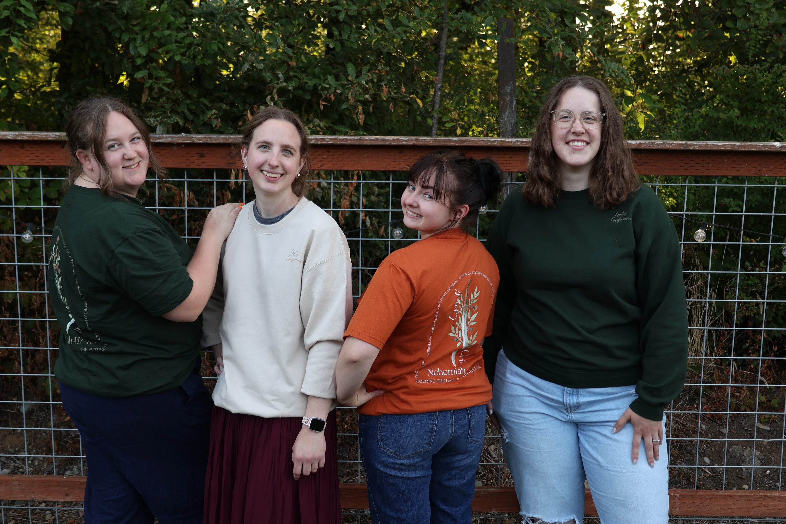 Four young women in casual attire stand together smiling for a photo in front of a wire fence with green trees in the background.