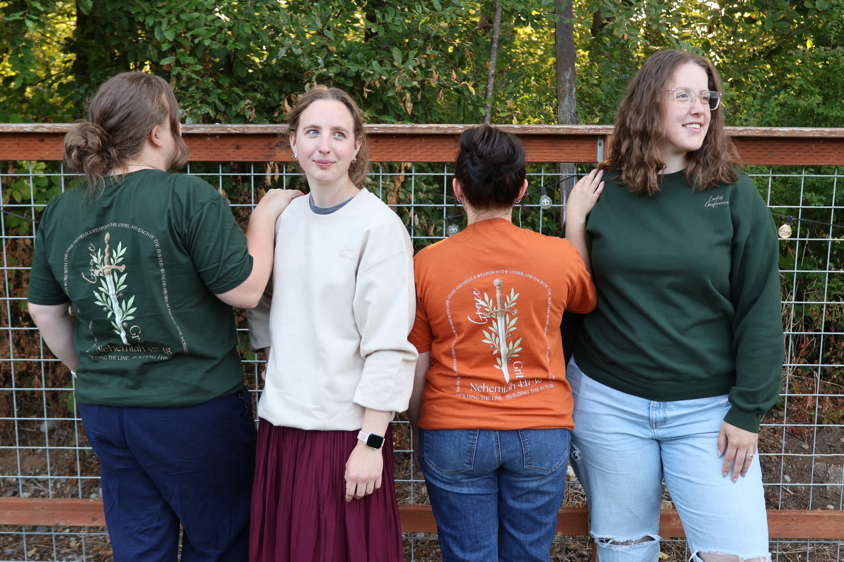 Four women stand together outdoors. Two face forward and two face backward, showing matching graphic t-shirts. They are in front of a fence and trees.