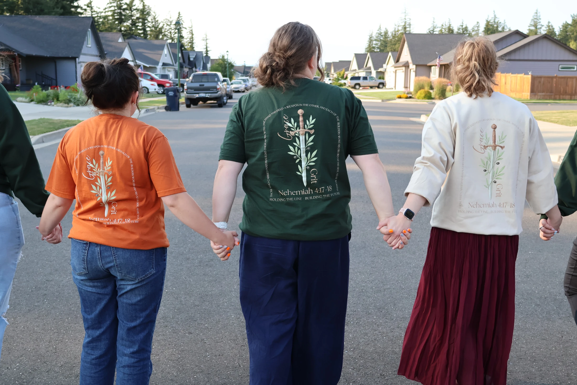 Three people seen from behind, holding hands in a line on a suburban street. They are wearing matching shirts with a sword and olive branch design.