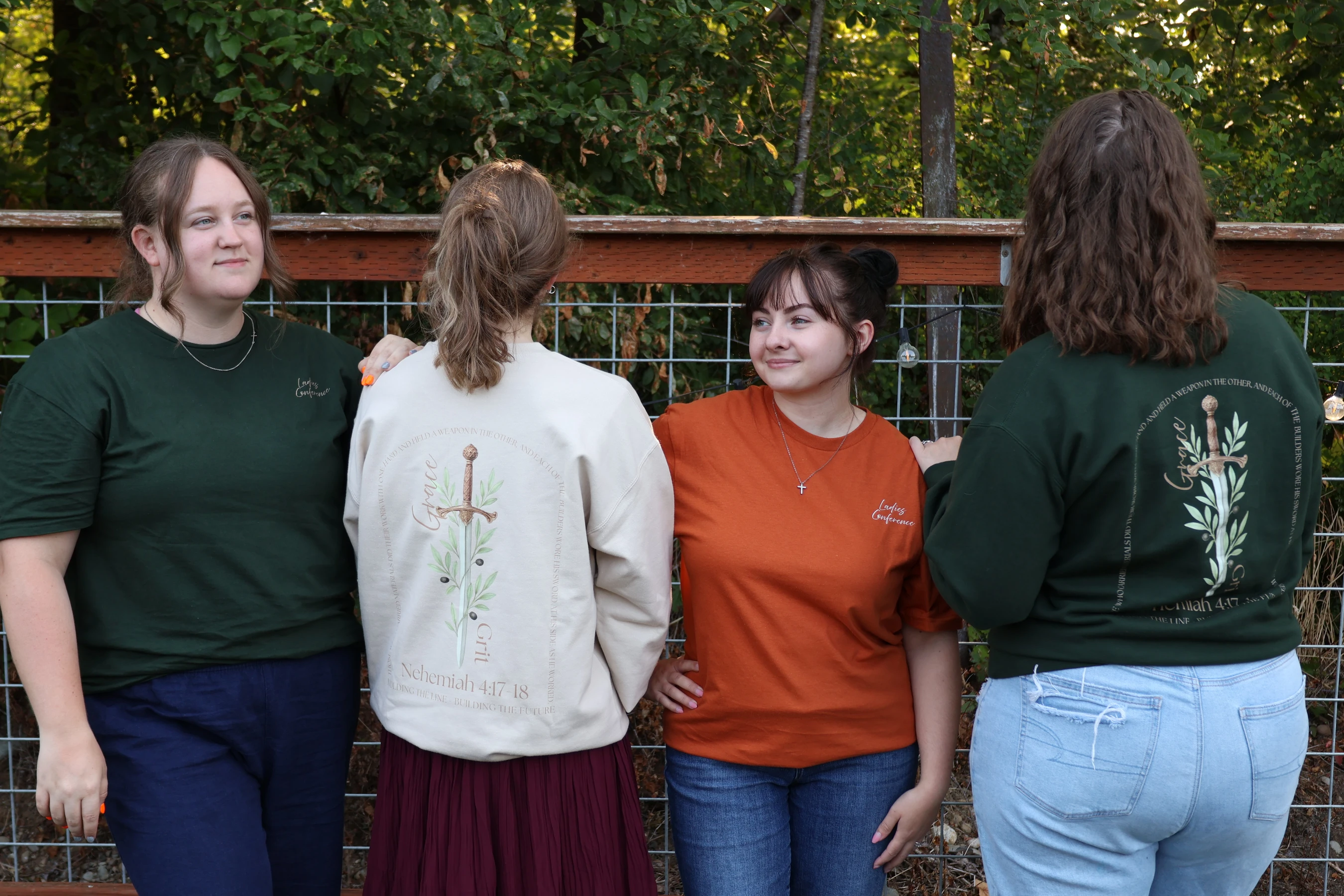 Four young women stand together outdoors. Two show the backs of sweatshirts with a sword and bible verse, while others wear conference t-shirts.