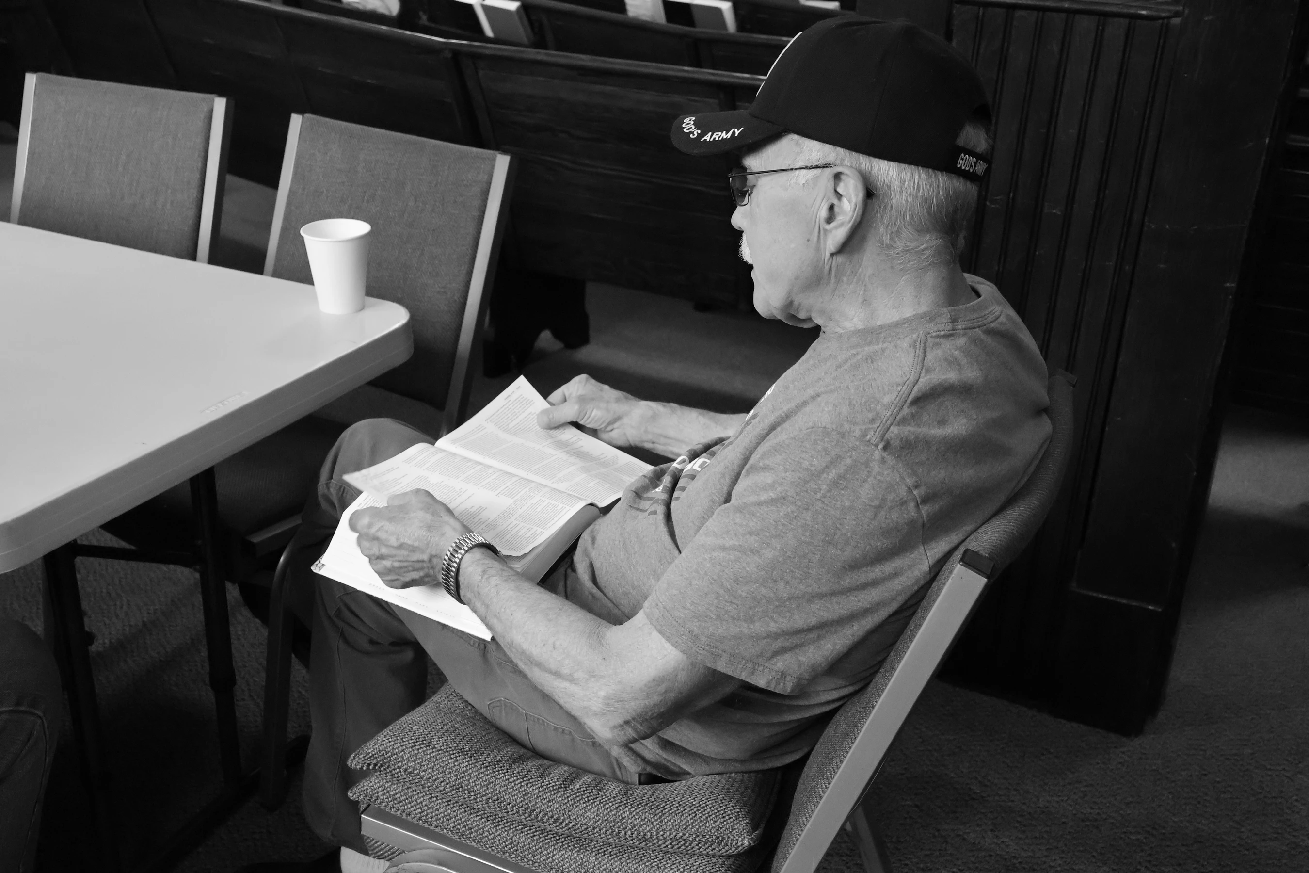 A black and white photo of an older man wearing a 'God's Army' cap sitting at a table in a church, intently reading a large open book, likely a Bible.