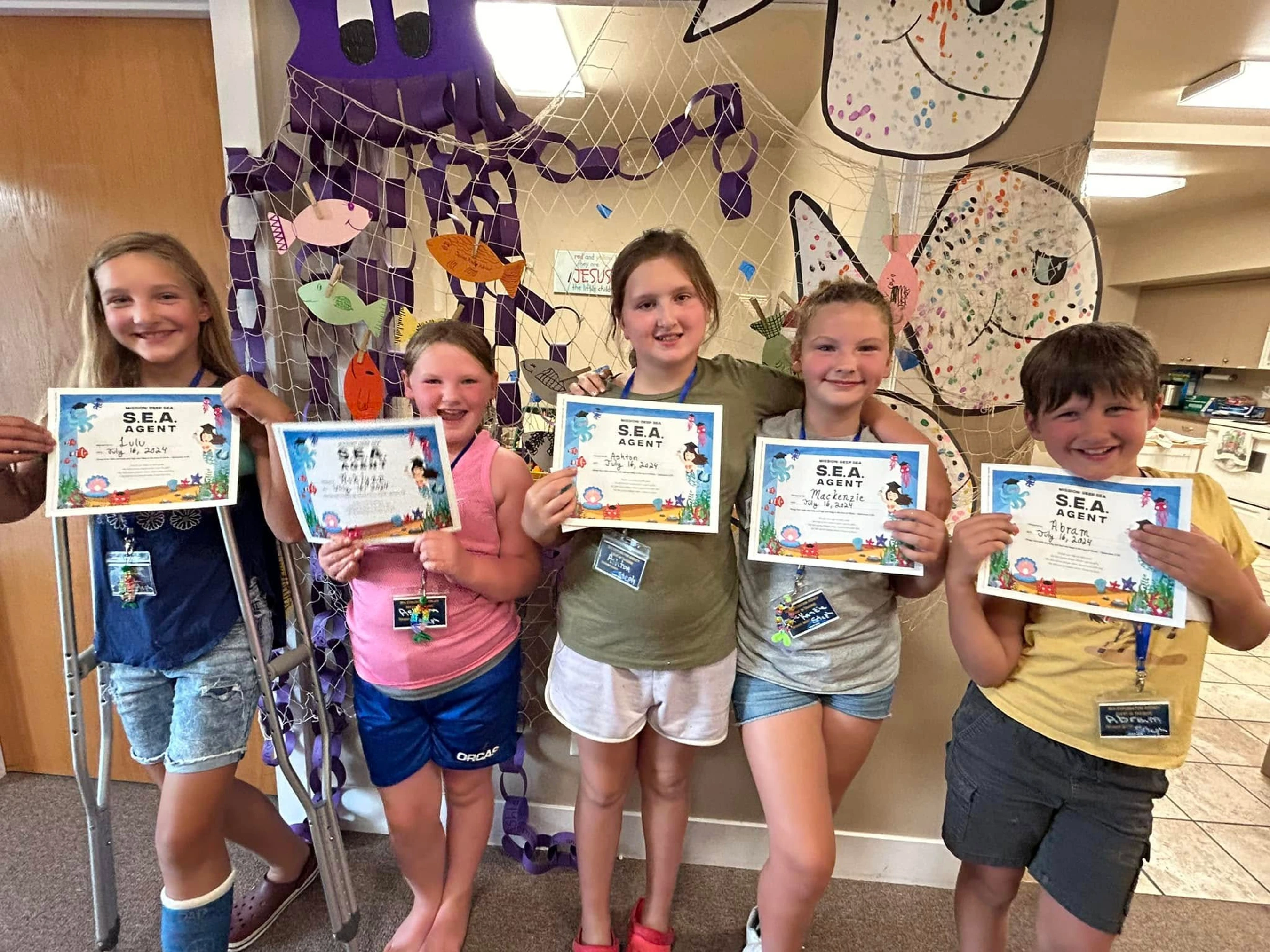 Five smiling children stand in front of an ocean-themed backdrop, holding up certificates designating them as 'S.E.A. Agents'.
