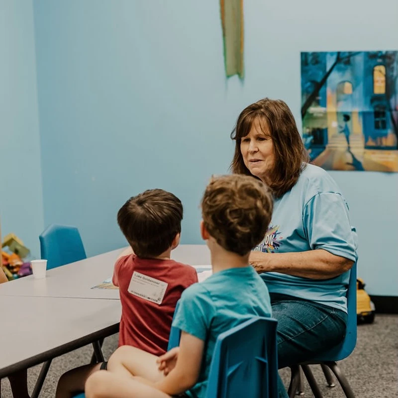 A woman with brown hair sits at a table and talks to two young boys whose backs are to the camera. They are in a classroom with light blue walls.