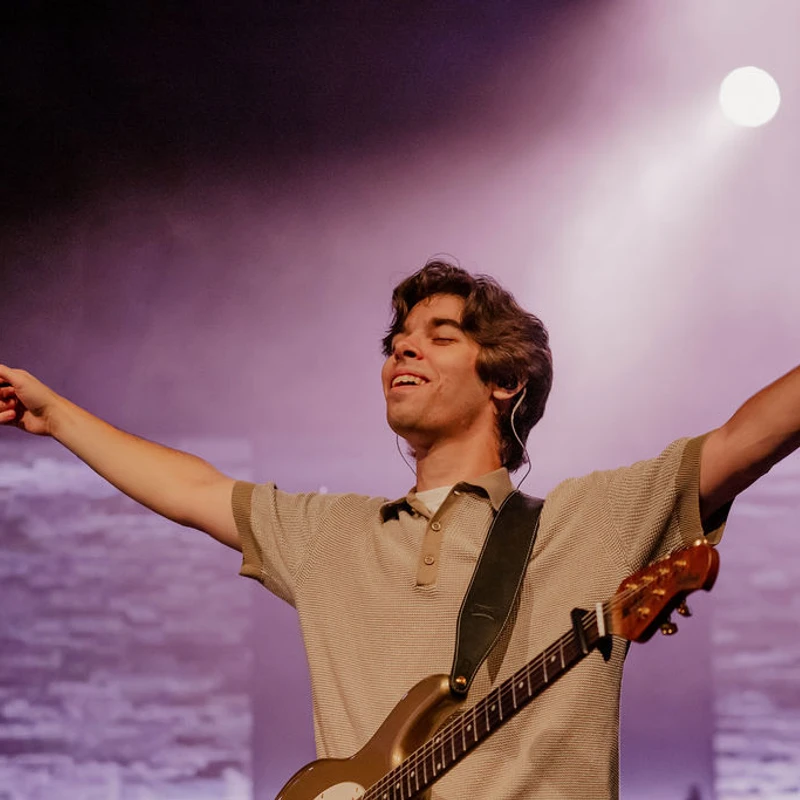 A young man with an electric guitar stands on stage with his arms outstretched, smiling with his eyes closed under a single bright spotlight.