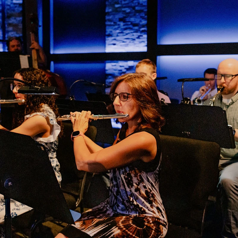 An orchestra performs on a stage with blue lighting. In the foreground, a woman in glasses plays a flute, next to a man playing a saxophone.