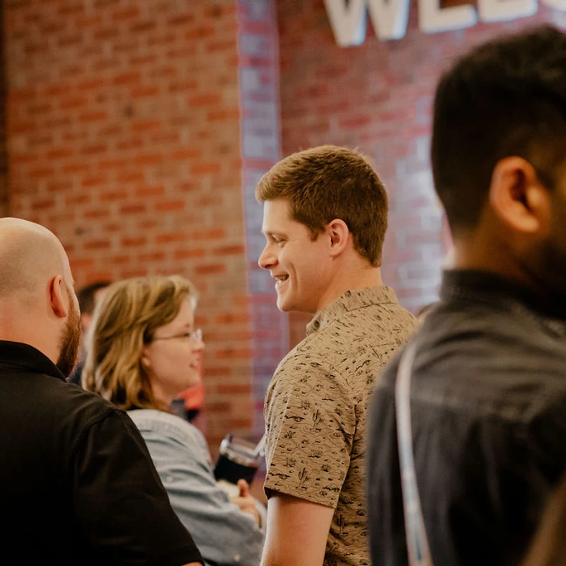 A man in a patterned shirt smiles while talking with others in a group at an indoor event. The setting has an exposed red brick wall.