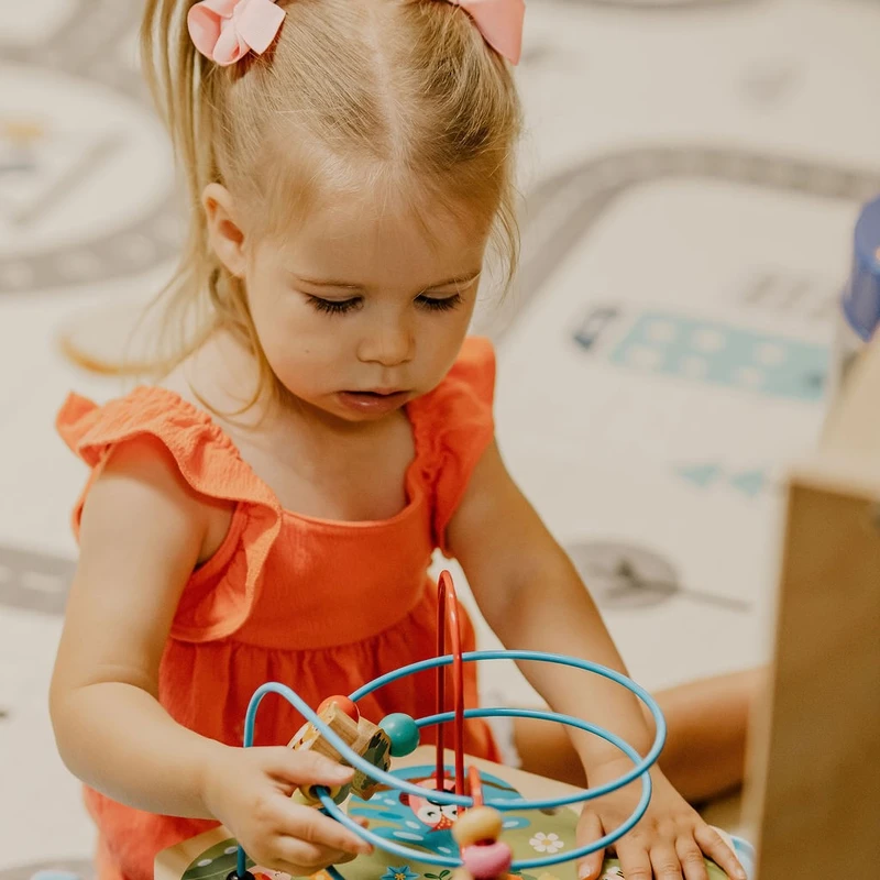 A young girl with blonde pigtails and pink bows concentrates on moving beads along the wires of a colorful wooden activity toy on a play mat.