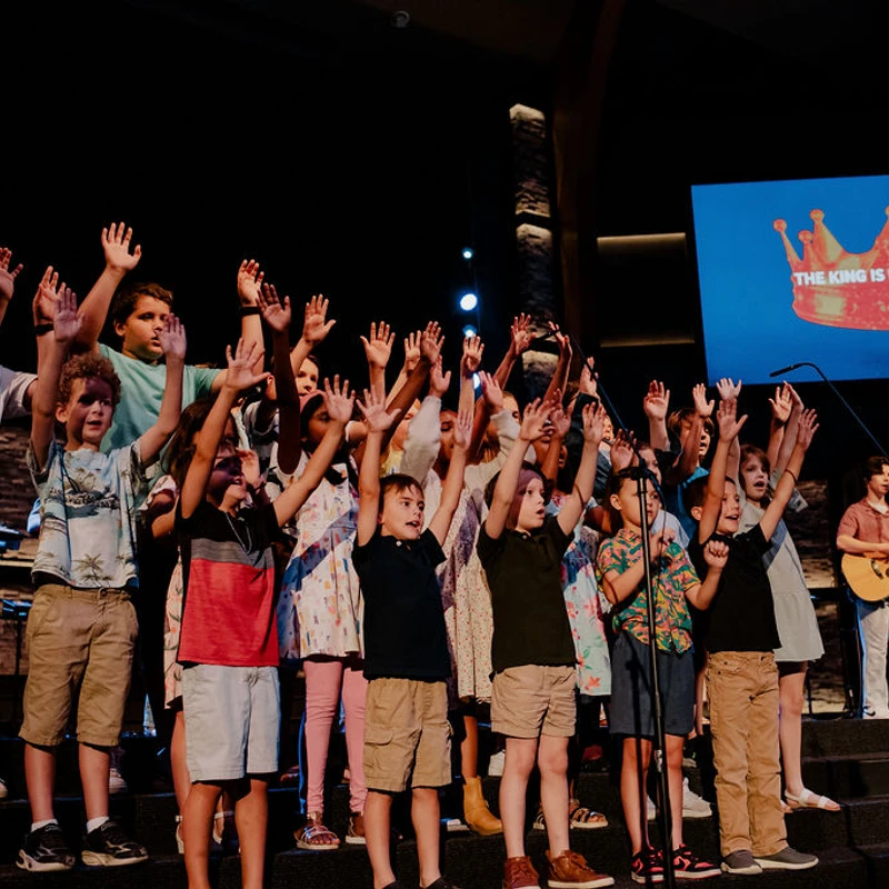 A large children's choir sings on stage with their hands raised. A screen behind them shows a crown and the words 'THE KING IS IN THE ROOM'.