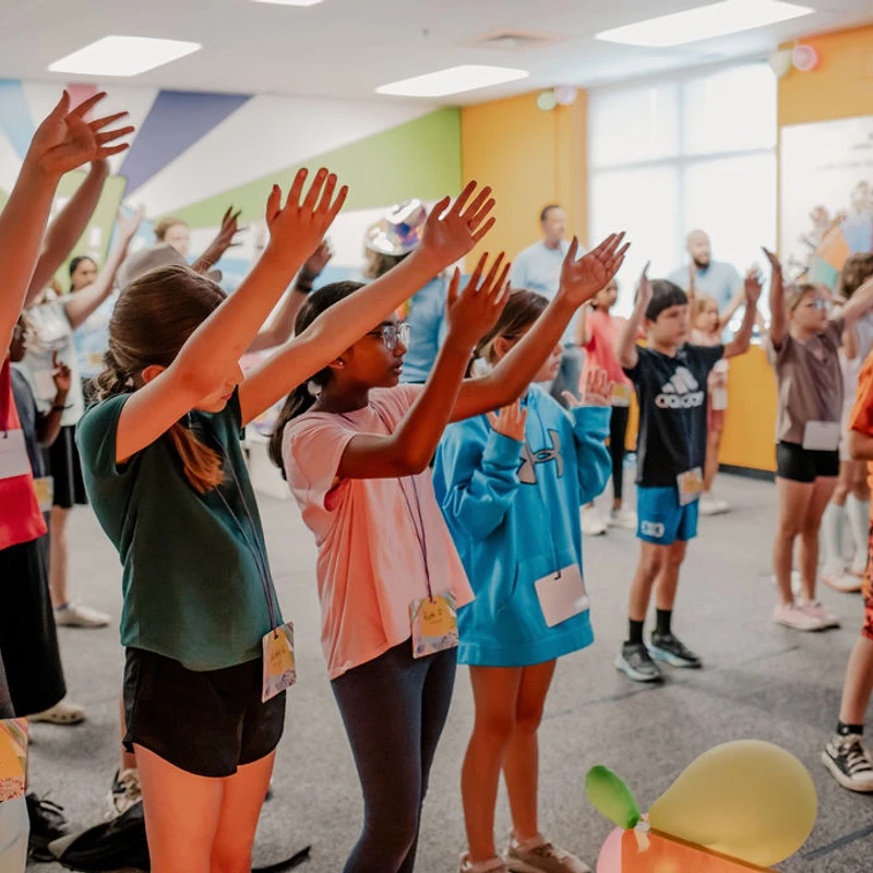A diverse group of elementary-aged children stand in a brightly colored room with their hands raised, enthusiastically participating in a group activity.