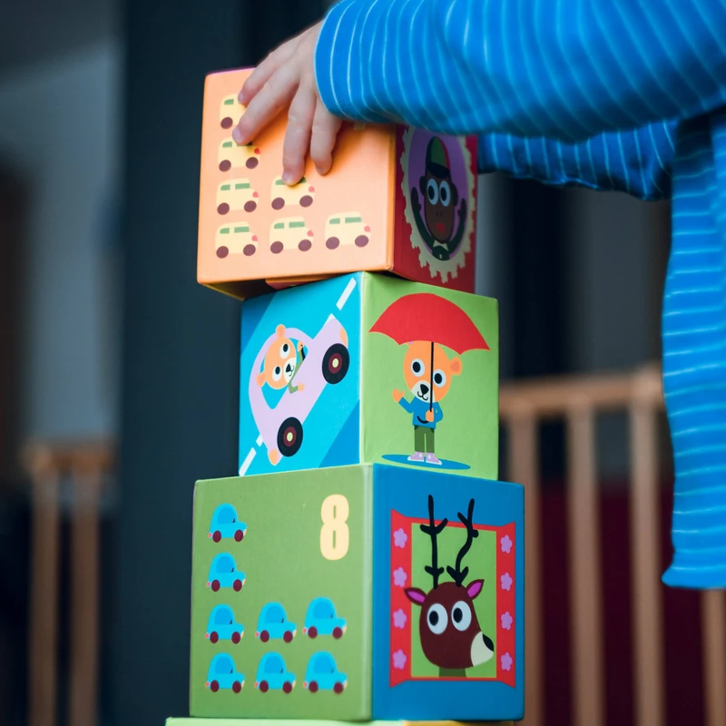 A child in a blue striped shirt stacks colorful toy blocks decorated with cartoon animals, cars, and numbers, building a tall tower.