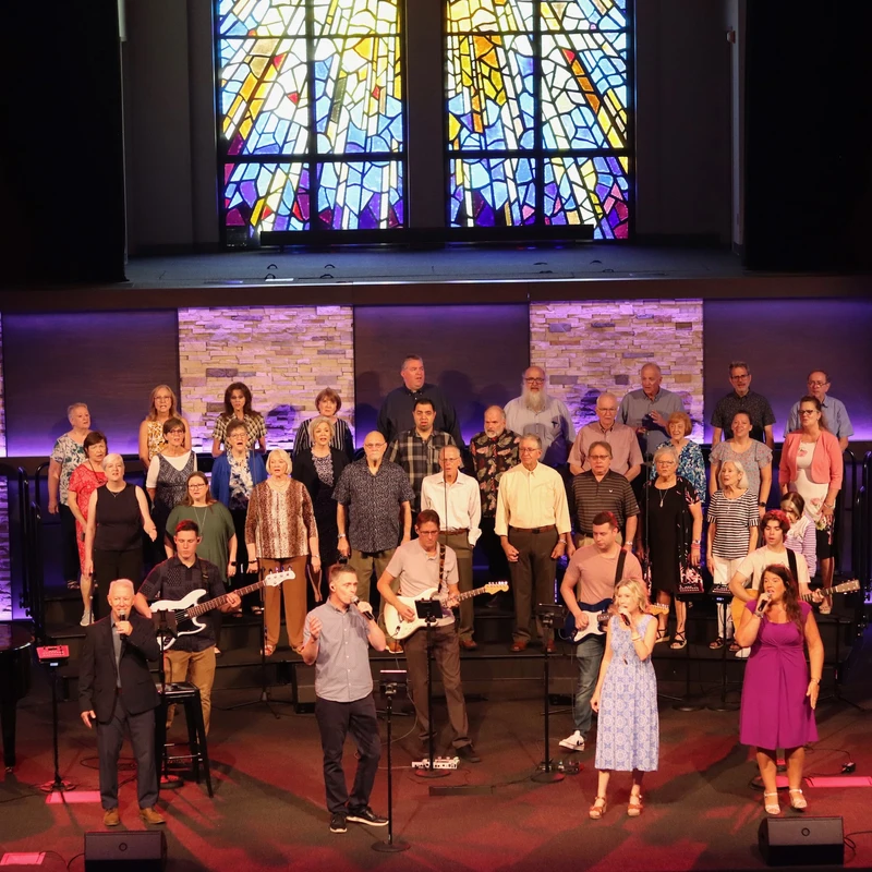 A large choir and worship band perform on a modern church stage with purple lighting. Vocalists sing at the front below a colorful stained-glass window.
