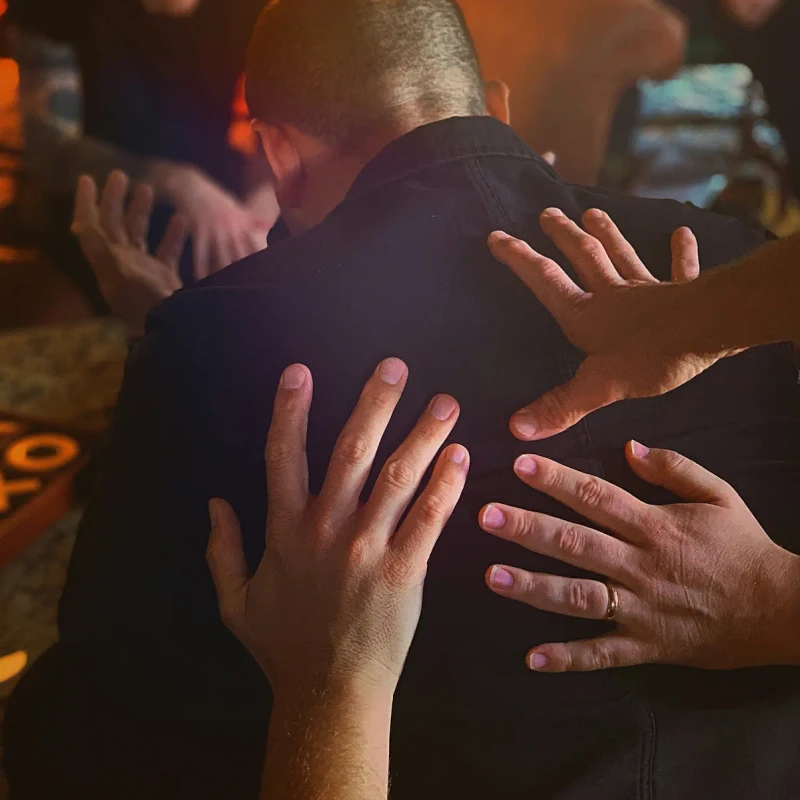 From behind, a man with a shaved head receives supportive touches on his back from several people in a group setting, suggesting prayer or comfort.