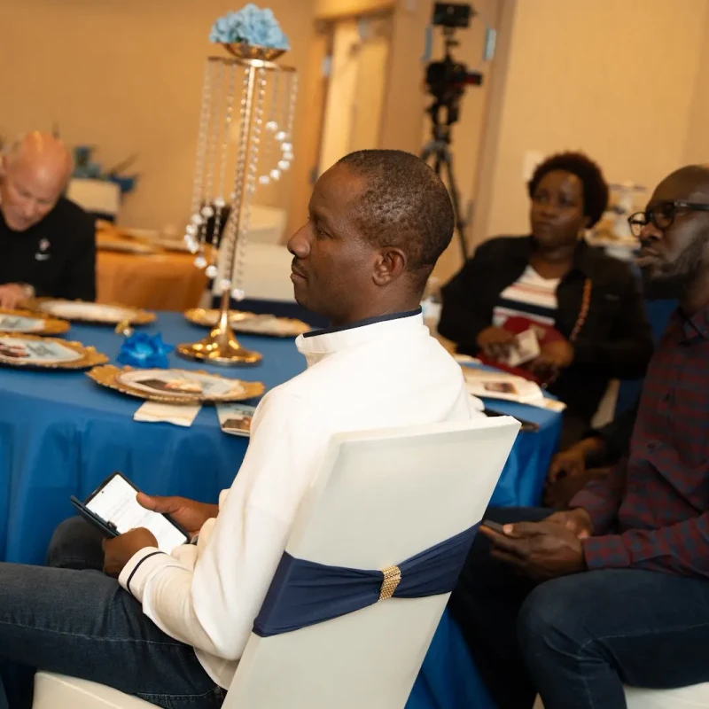Several people seated at decorated tables in a conference hall. A man in a white shirt in the foreground holds a phone while listening with others.