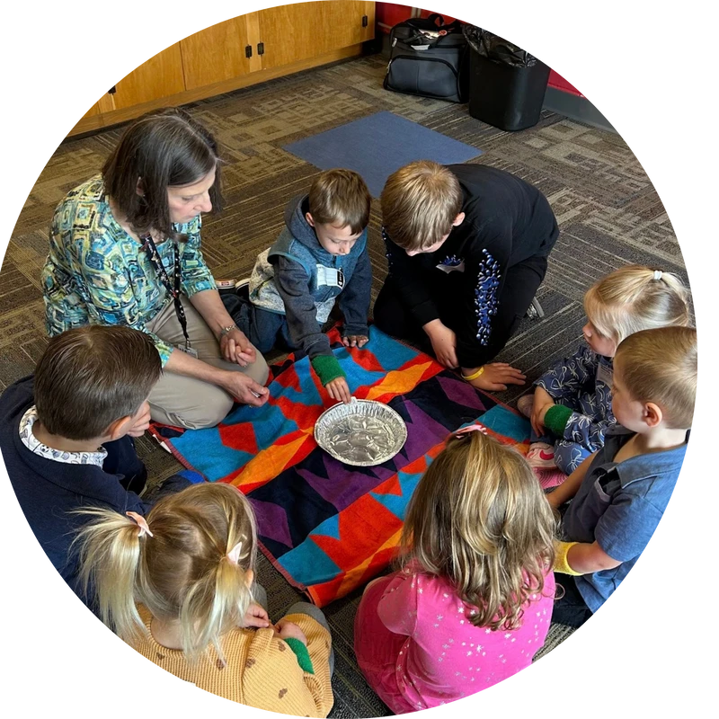 A female teacher and a group of young children sit in a circle on a classroom floor, focused on a hands-on learning activity with a foil pan.