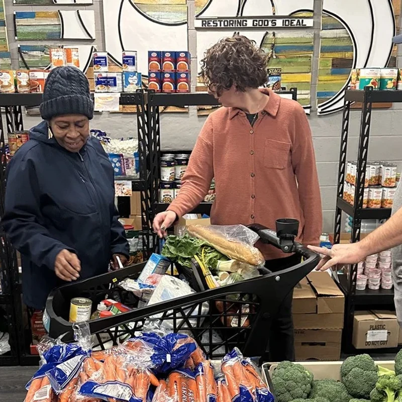 Three people stand around a shopping cart filled with fresh produce and groceries inside a food pantry with shelves stocked with canned and boxed goods.