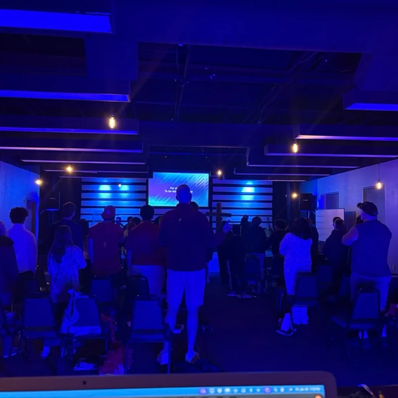 View from the back of a room where a congregation worships under blue lights, facing a stage with a screen displaying text.