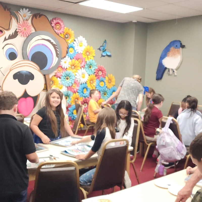 A group of children and a few adults gathered at tables for an art activity in a room with a large, colorful mural of a cartoon dog and paper flowers.