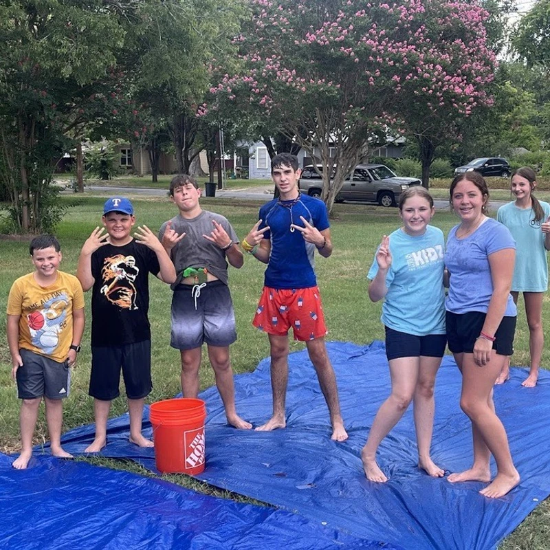 A group of seven smiling kids and teens in shorts and t-shirts stand on a wet blue tarp on a lawn, posing for a photo after playing on a water slide.