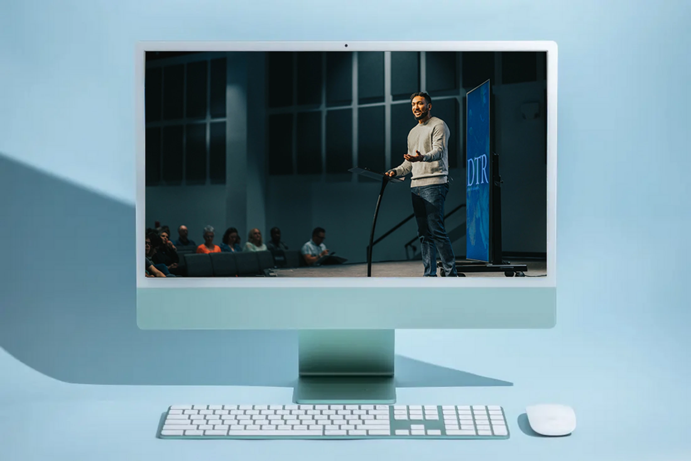 A desktop computer on a blue background displays a video of a man speaking at a podium to a live audience, illustrating a virtual conference or online event.