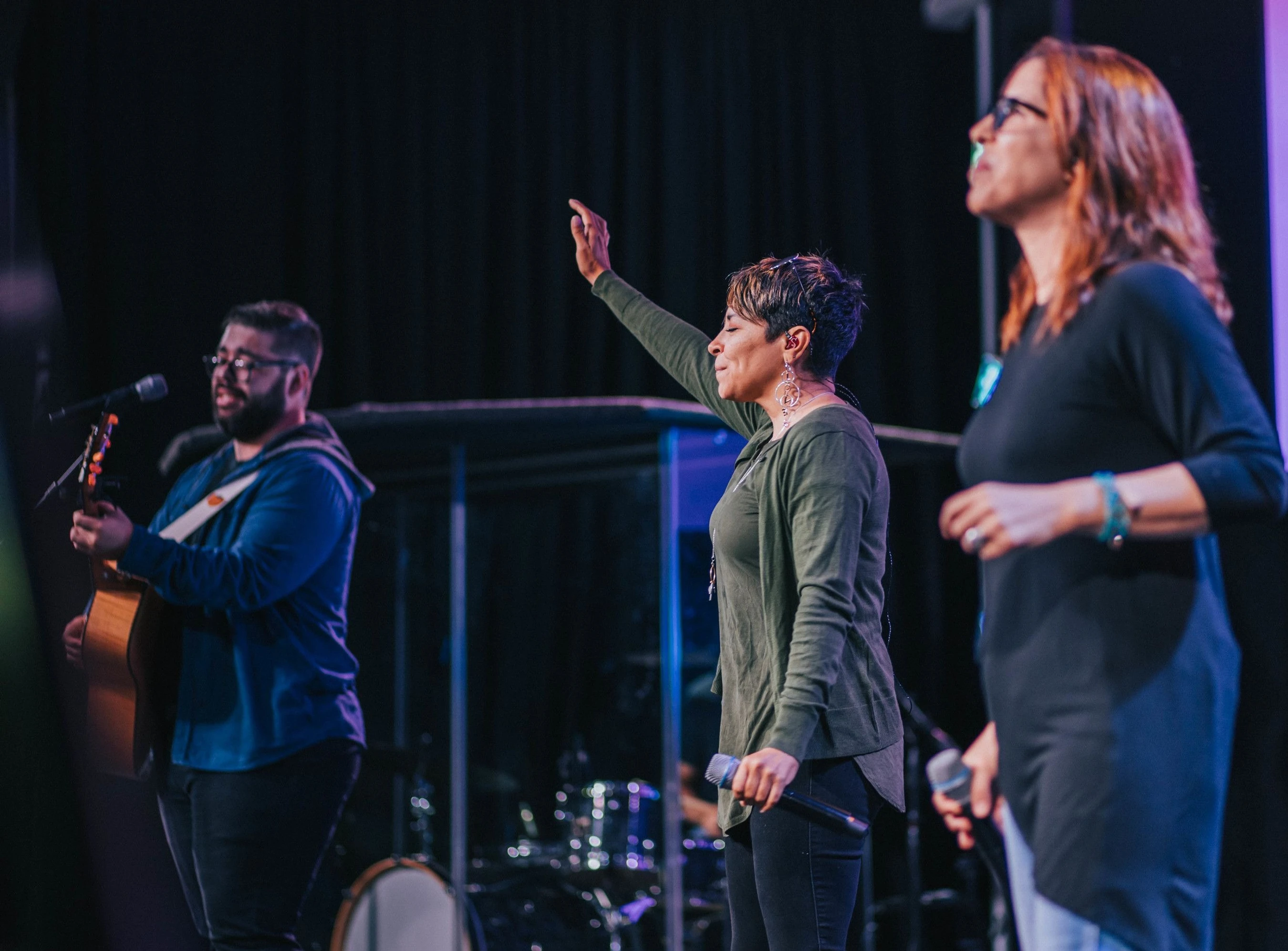 A band performs on stage. A man plays guitar while two women sing. The woman in the center has her hand raised and eyes closed in worship.