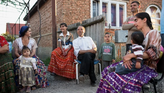 A large family with several generations, including children and an elder, gathered outdoors in front of a brick house, wearing colorful traditional clothing.