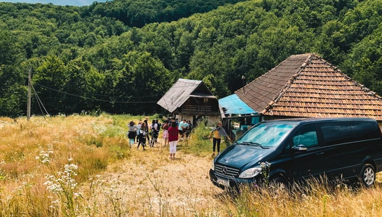 A black minivan is parked in a grassy field near two rustic houses. A group of people walks nearby, with a large, forest-covered hill in the background.