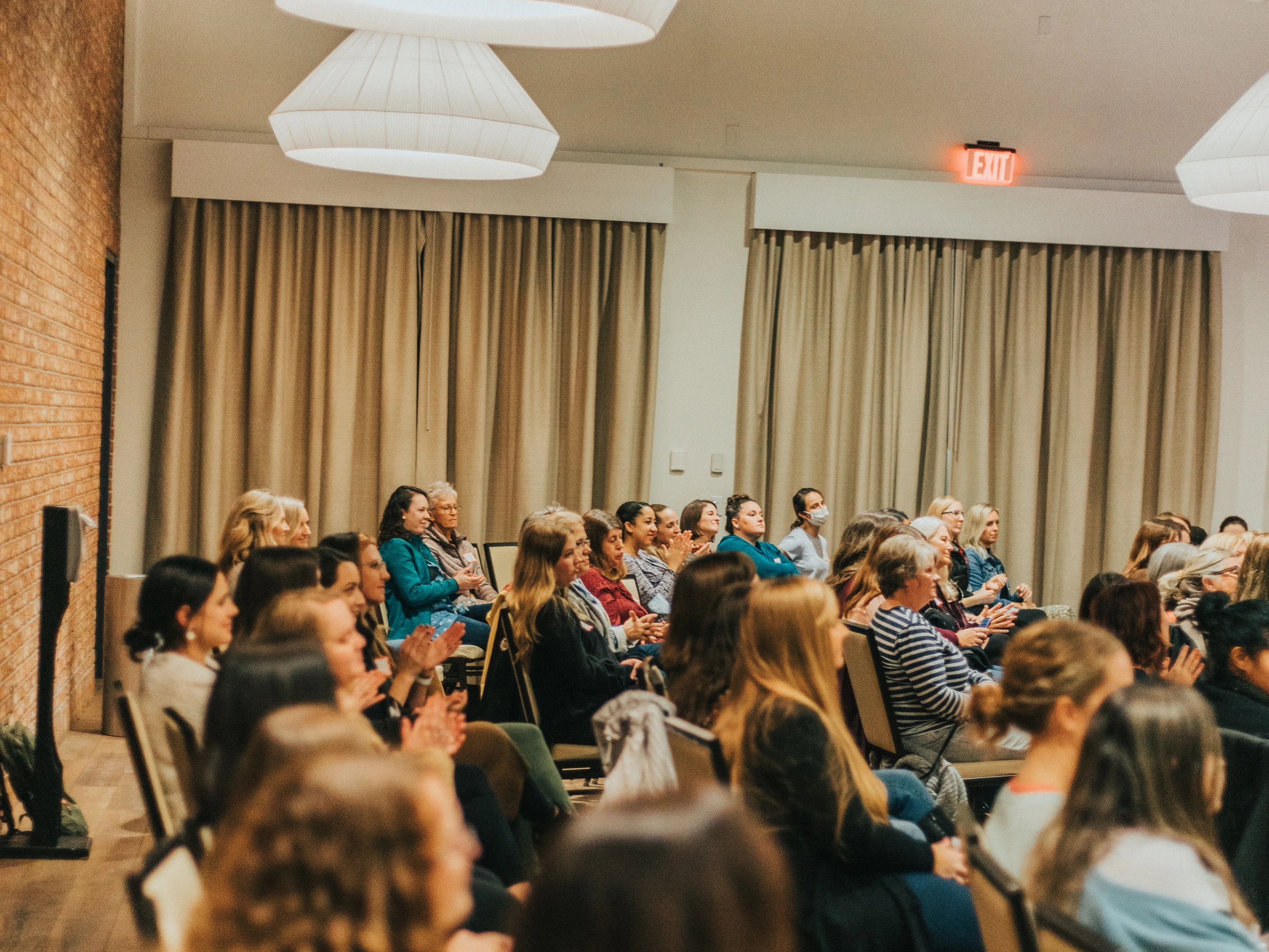 An audience of mostly women sit in chairs in a conference room, listening attentively to a presentation off-camera. Some people are clapping.