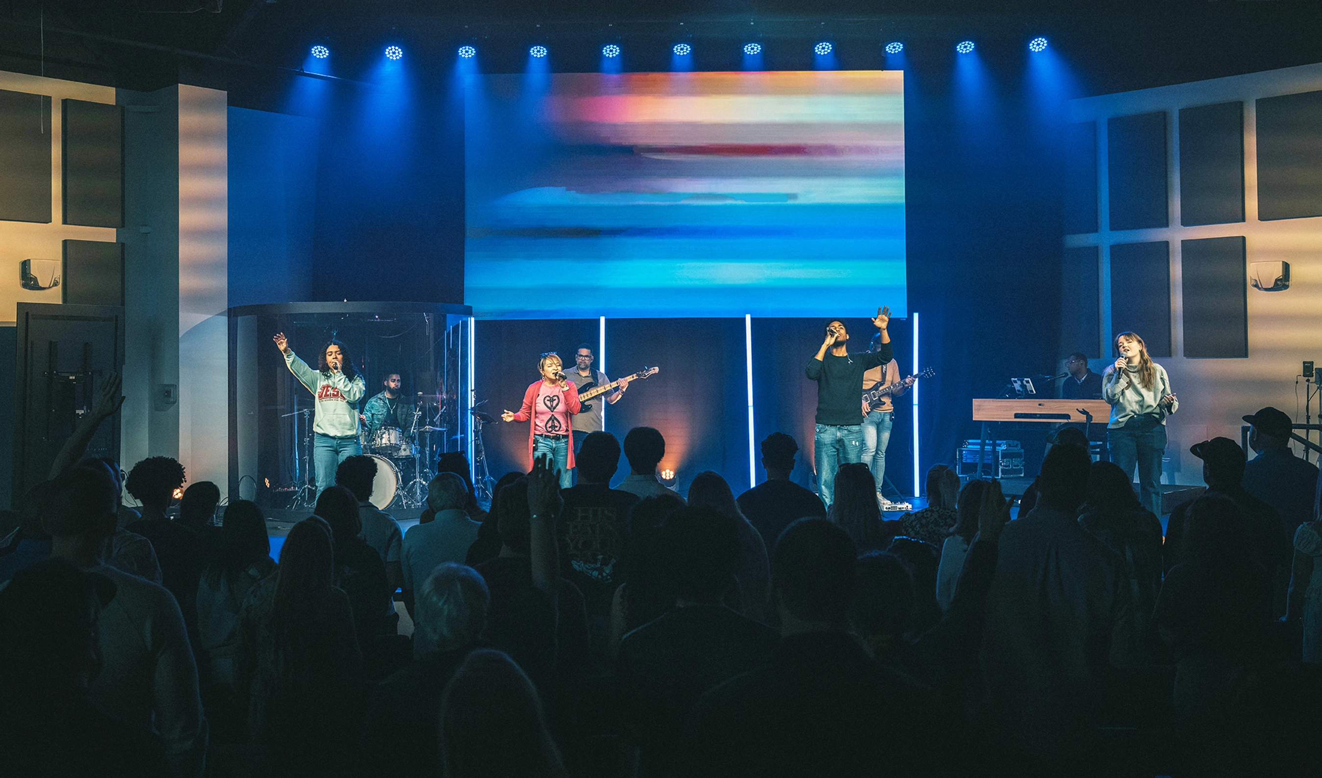 A band with several singers and musicians performs on a modern stage with a large screen for a large audience, all under cool blue stage lights.