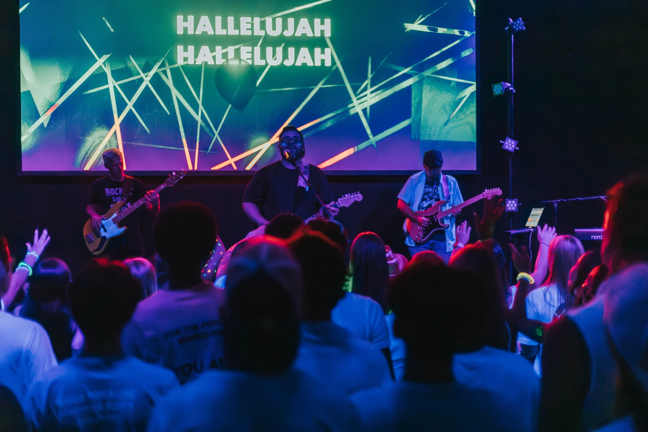 A Christian worship band with three guitarists performs on stage. The crowd below raises their hands as a screen in the background displays 'Hallelujah'.