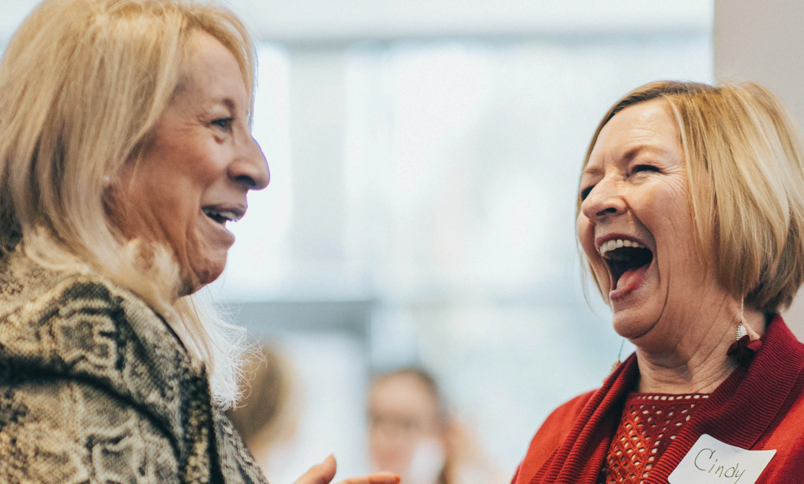 A close-up of two older blonde women laughing during a conversation. The woman on the right laughs with her mouth open, wearing a red sweater and a nametag.