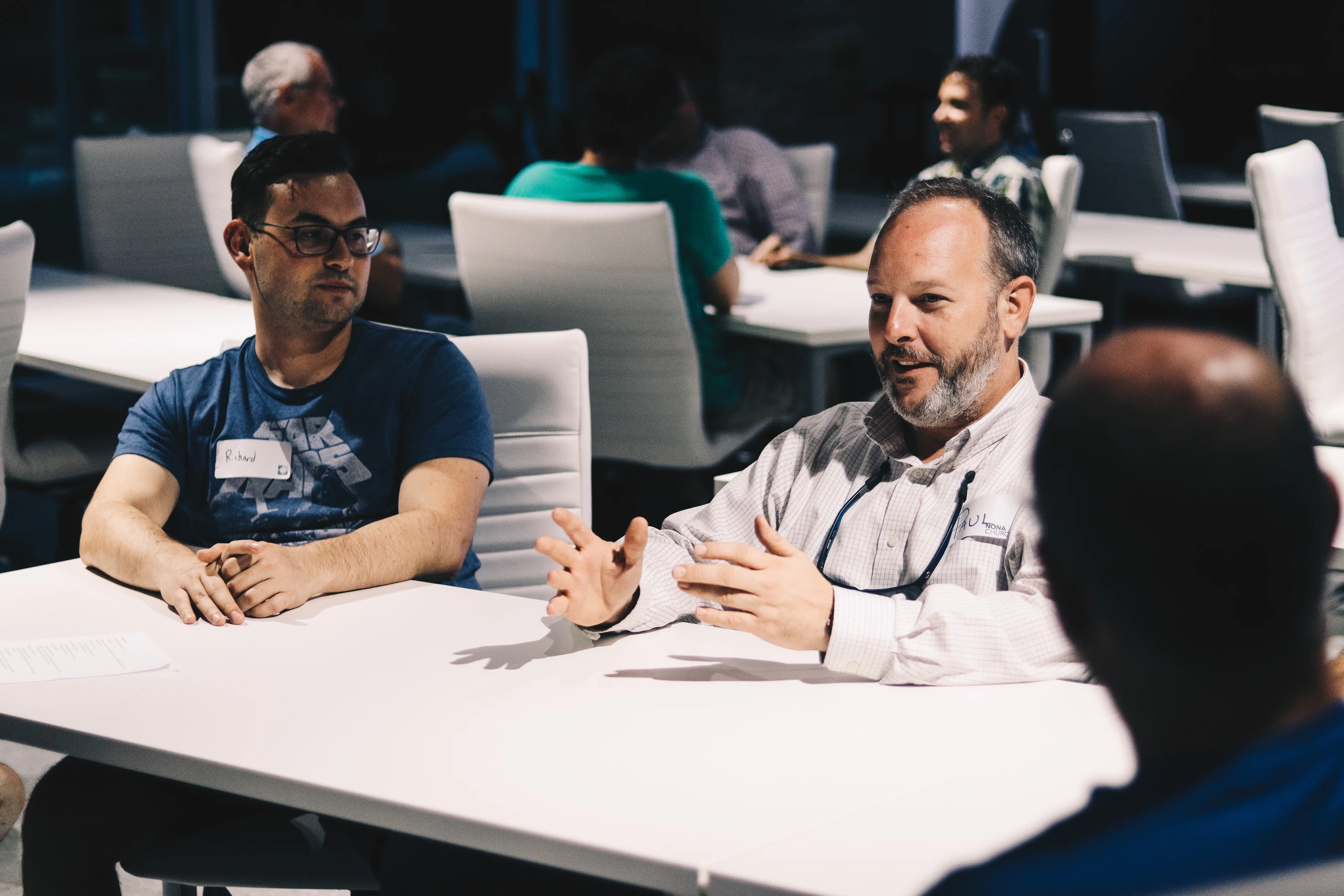 A man with a beard gestures as he speaks to two other men at a white table during a meeting, while the man next to him listens intently.