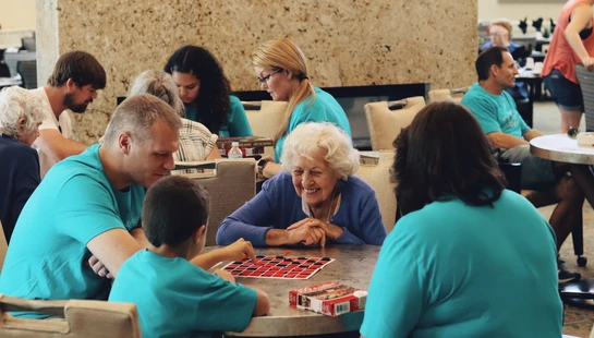 An elderly woman with white hair smiles as she plays a game of checkers with a young boy and his family, who are all wearing matching turquoise shirts.
