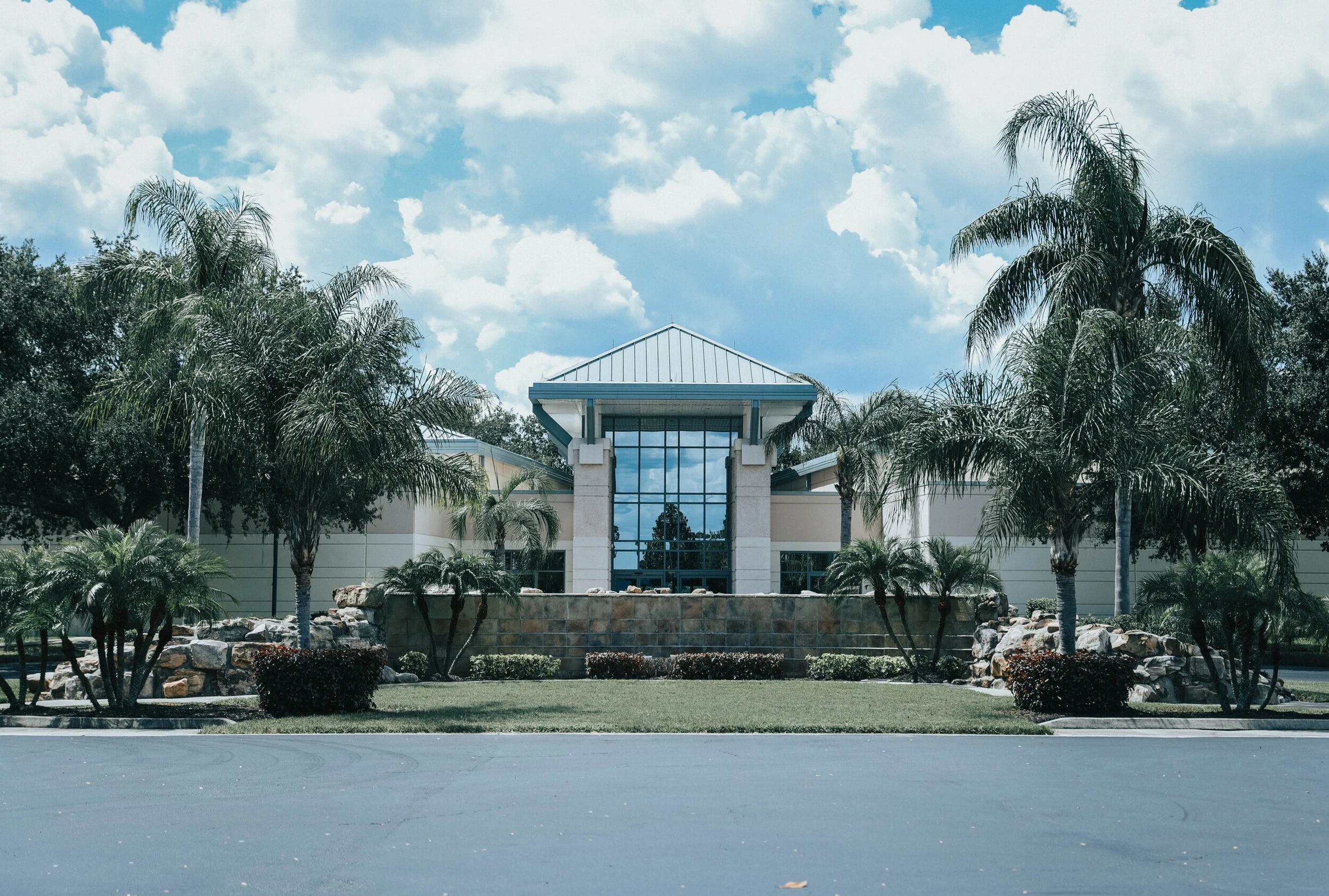 Front view of a modern building with a glass facade, flanked by tall palm trees and lush landscaping under a partly cloudy blue sky.