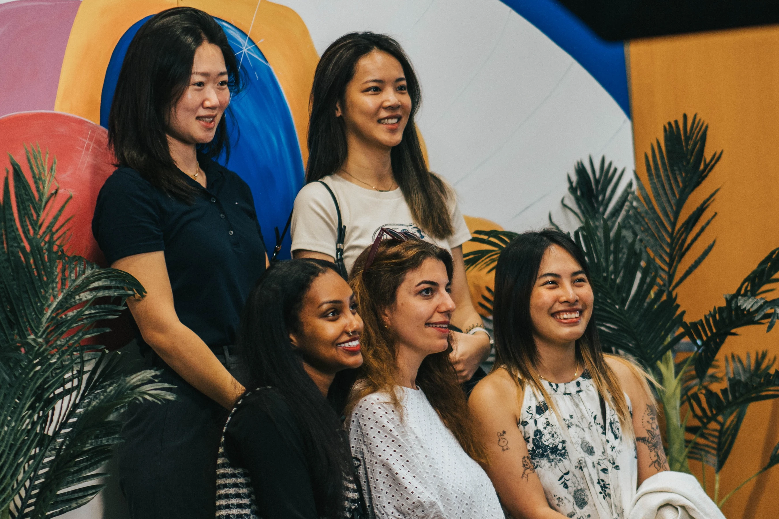 A diverse group of five smiling women posing for a photo together against a colorful, abstract background with some green plant leaves.