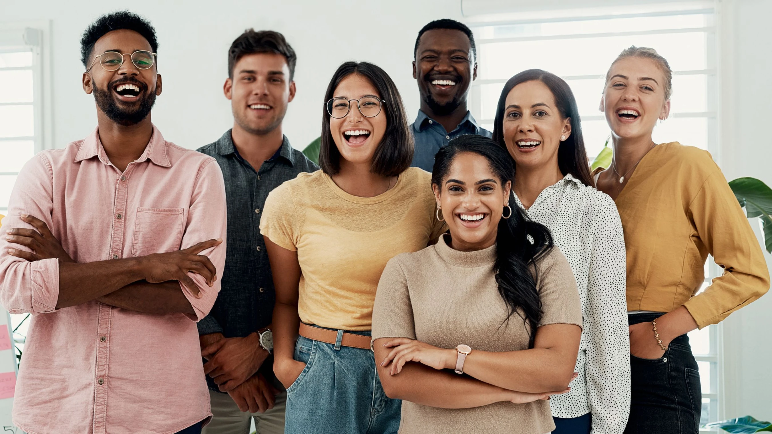 A group portrait of seven diverse and happy colleagues standing together in a bright office, smiling and laughing at the camera.