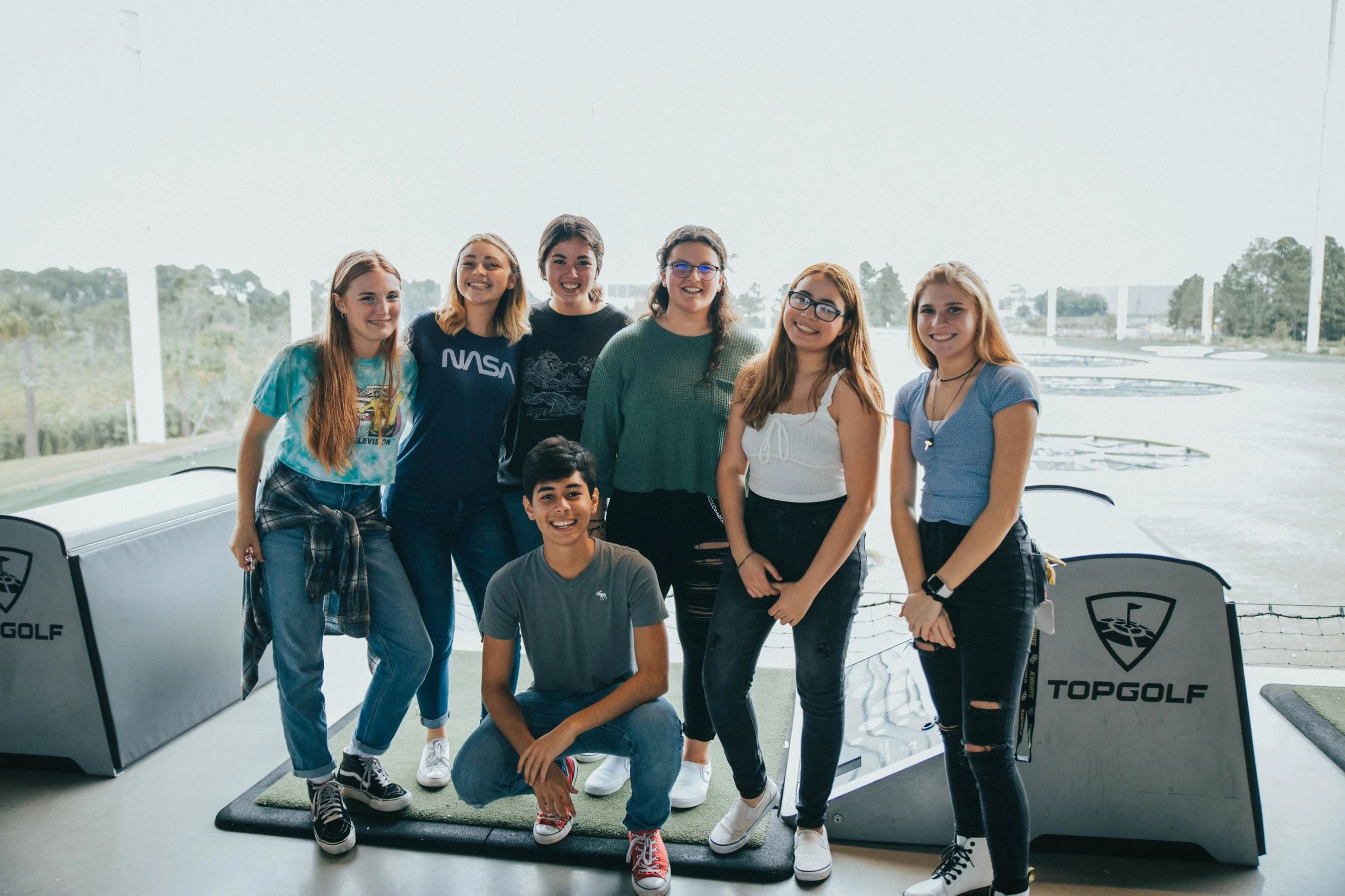 A group of seven teenagers, six girls standing and one boy crouching in front, smiling for a photo inside a Topgolf driving range bay.