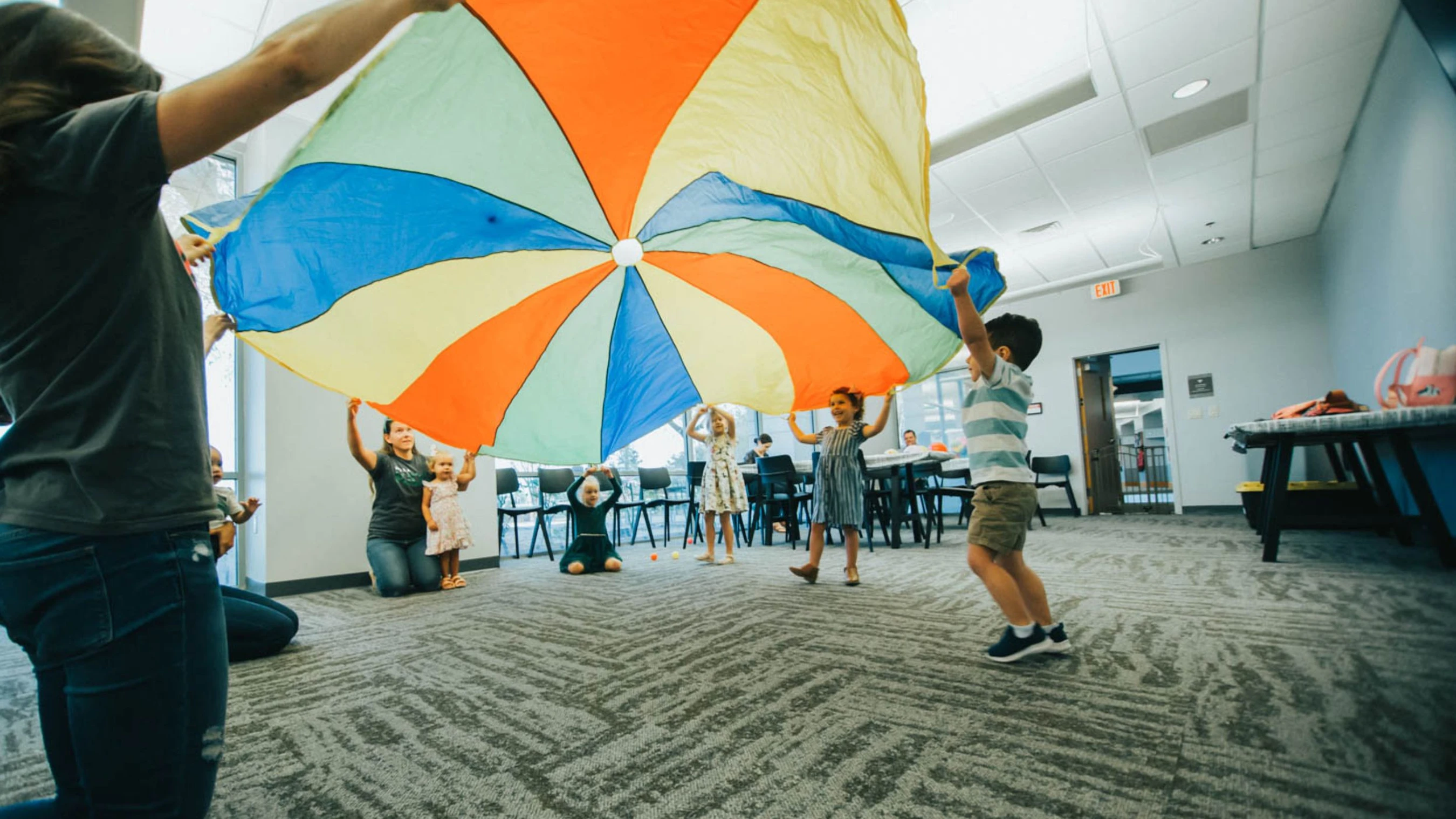 A group of children and adults play indoors, holding up a large, multicolored parachute in a community room with patterned gray carpet.