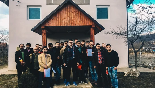 A group of about twenty men pose for a photo on the steps of a small, white church building under a bright blue sky.