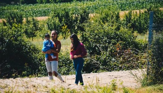 A woman holds a small child while another young woman stands beside them on a dirt path next to a lush green field on a sunny day.