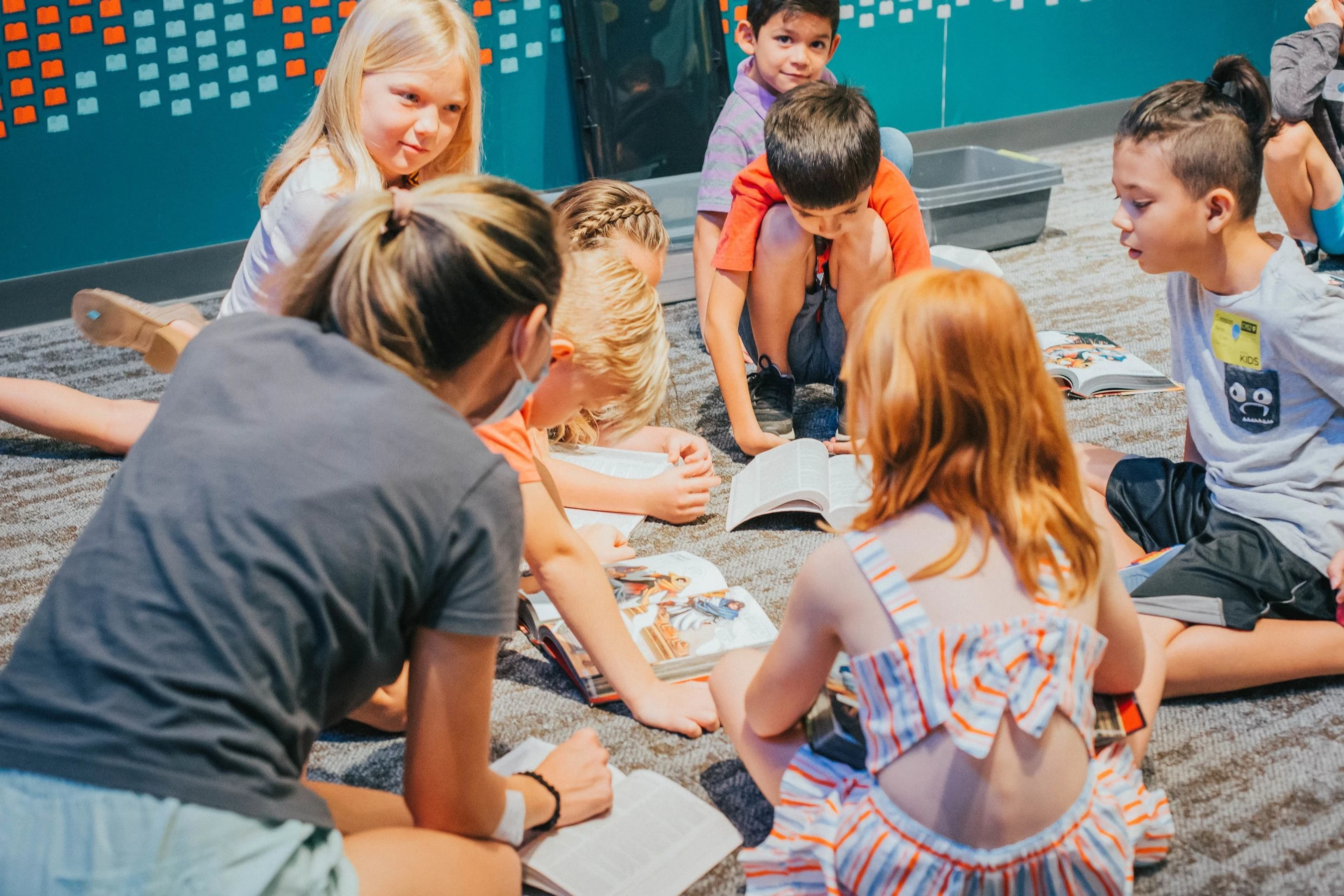 A group of diverse elementary-aged children and an adult sit in a circle on a carpeted floor, attentively reading books together in a classroom setting.