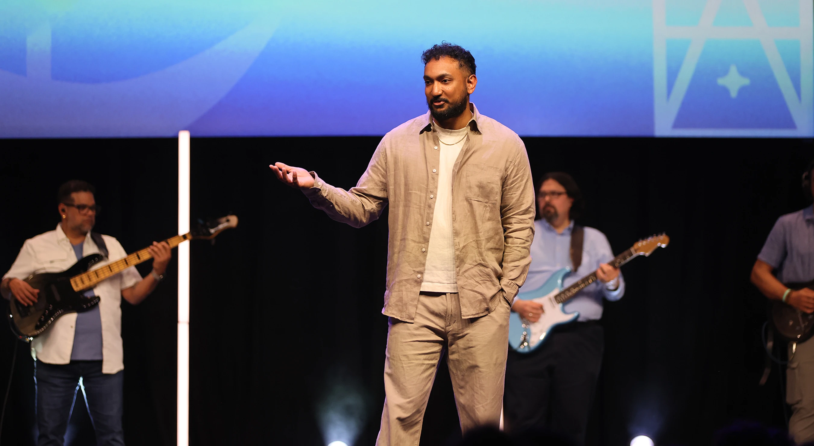 A man in a tan outfit stands on a stage speaking, with one hand gesturing outwards. In the background, a band with guitarists plays in front of a blue screen.