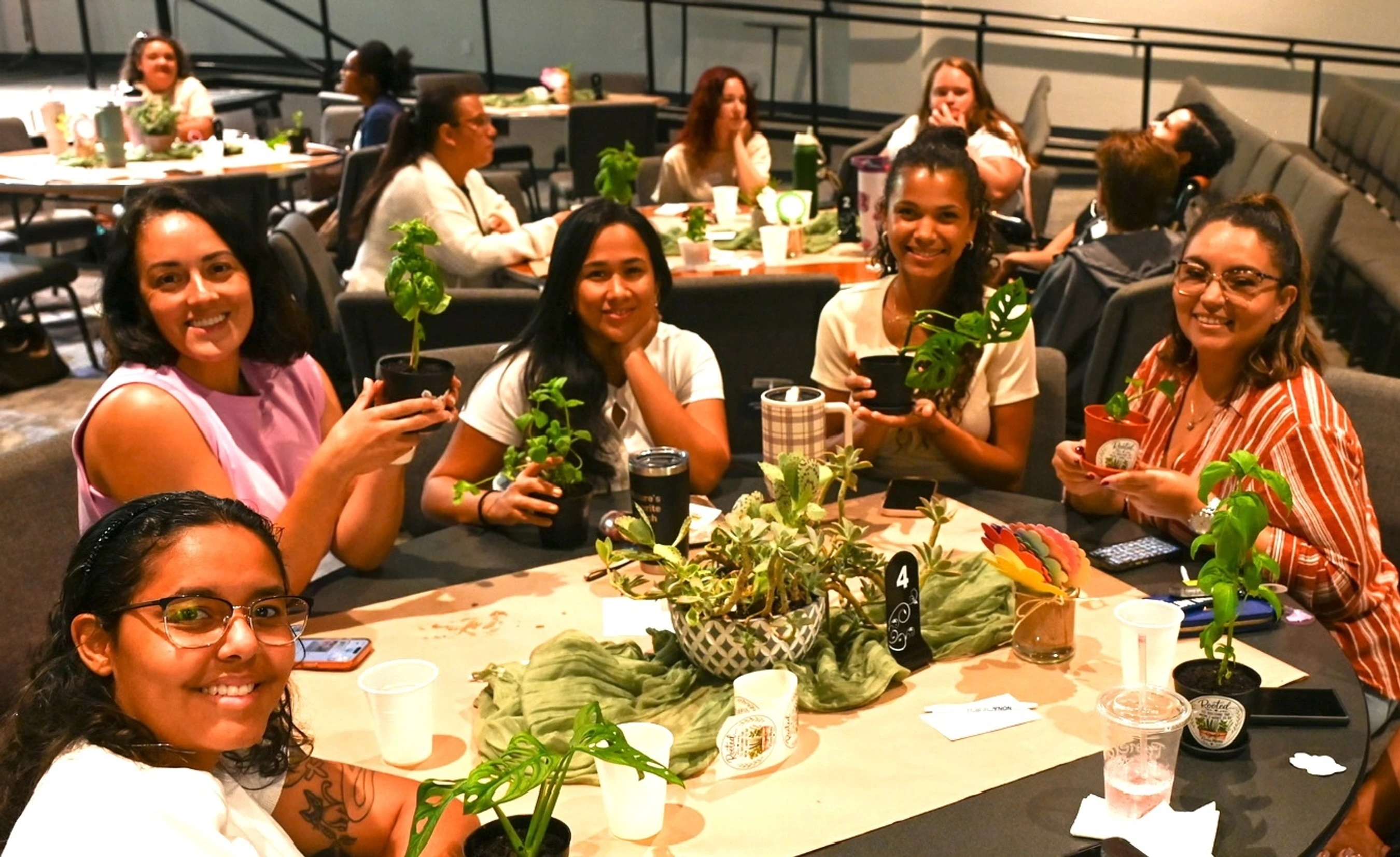 A group of five women smile for the camera while sitting at a round table during an event, each holding a small potted plant. The table has a plant centerpiece.