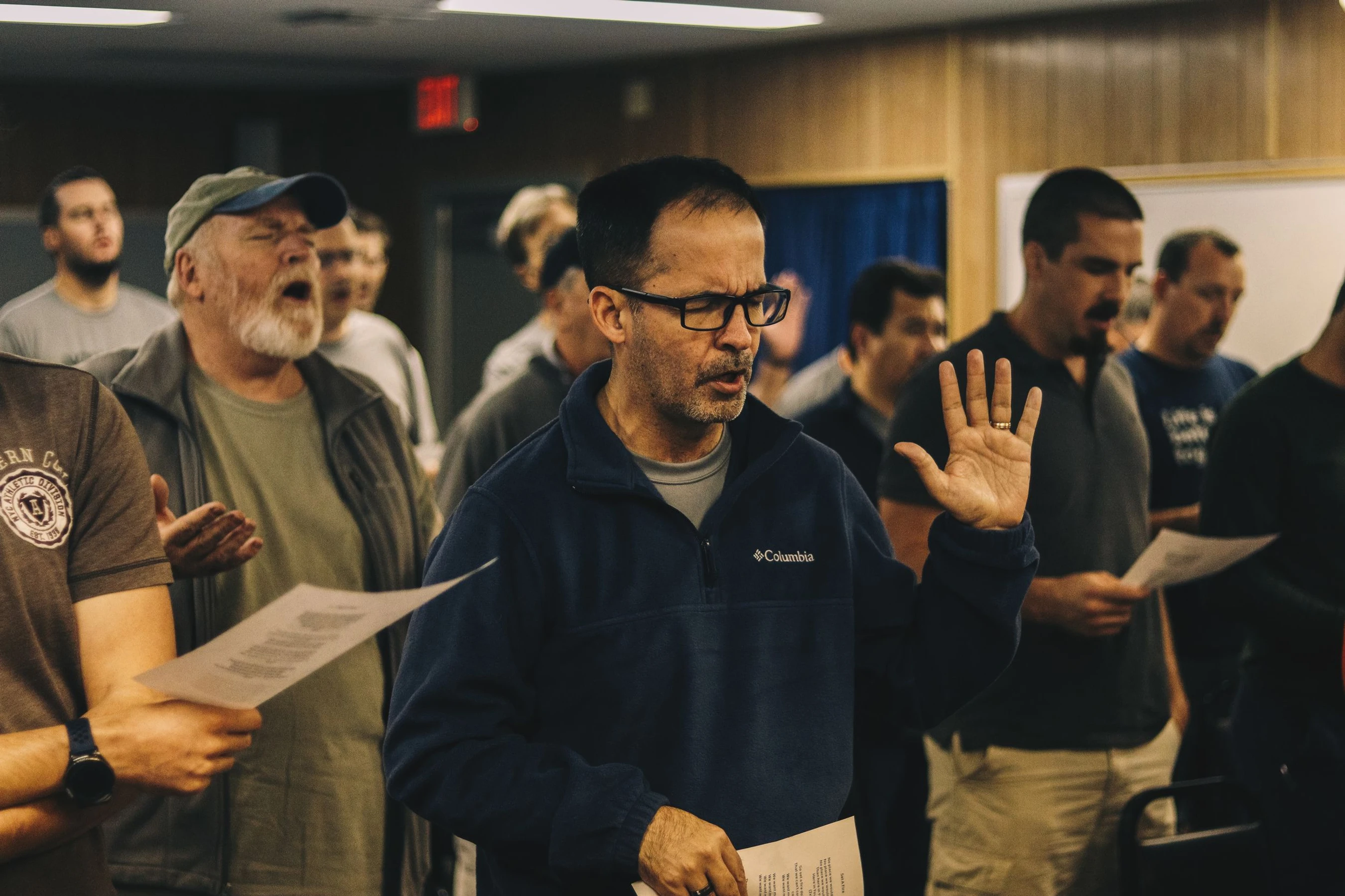 A group of men singing together indoors. One man in a blue fleece raises his hand while another with a white beard sings with his eyes closed.