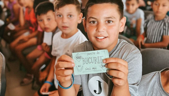 A young boy in a grey shirt smiles at the camera while holding up a small green ticket. He is sitting in a room with other children in the background.