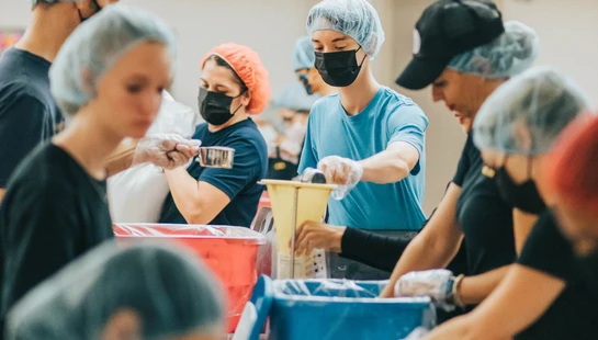 A group of volunteers wearing hairnets, gloves, and face masks work together to pack food items at an assembly line with large colorful bins.